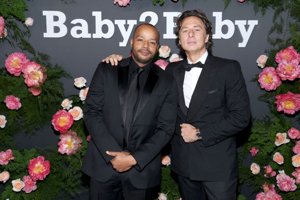 Zach Braff and Donald Faison dressed in suits smiling together at an event. They are surrounded by flowers, and Zac has his arm on Donald's shoulder